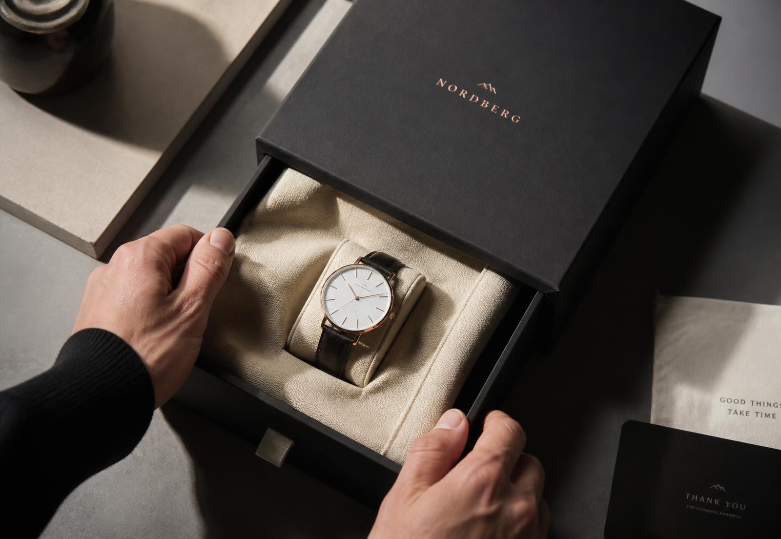 Hands opening a charcoal rigid drawer box revealing a luxury watch on a beige silk cushion
